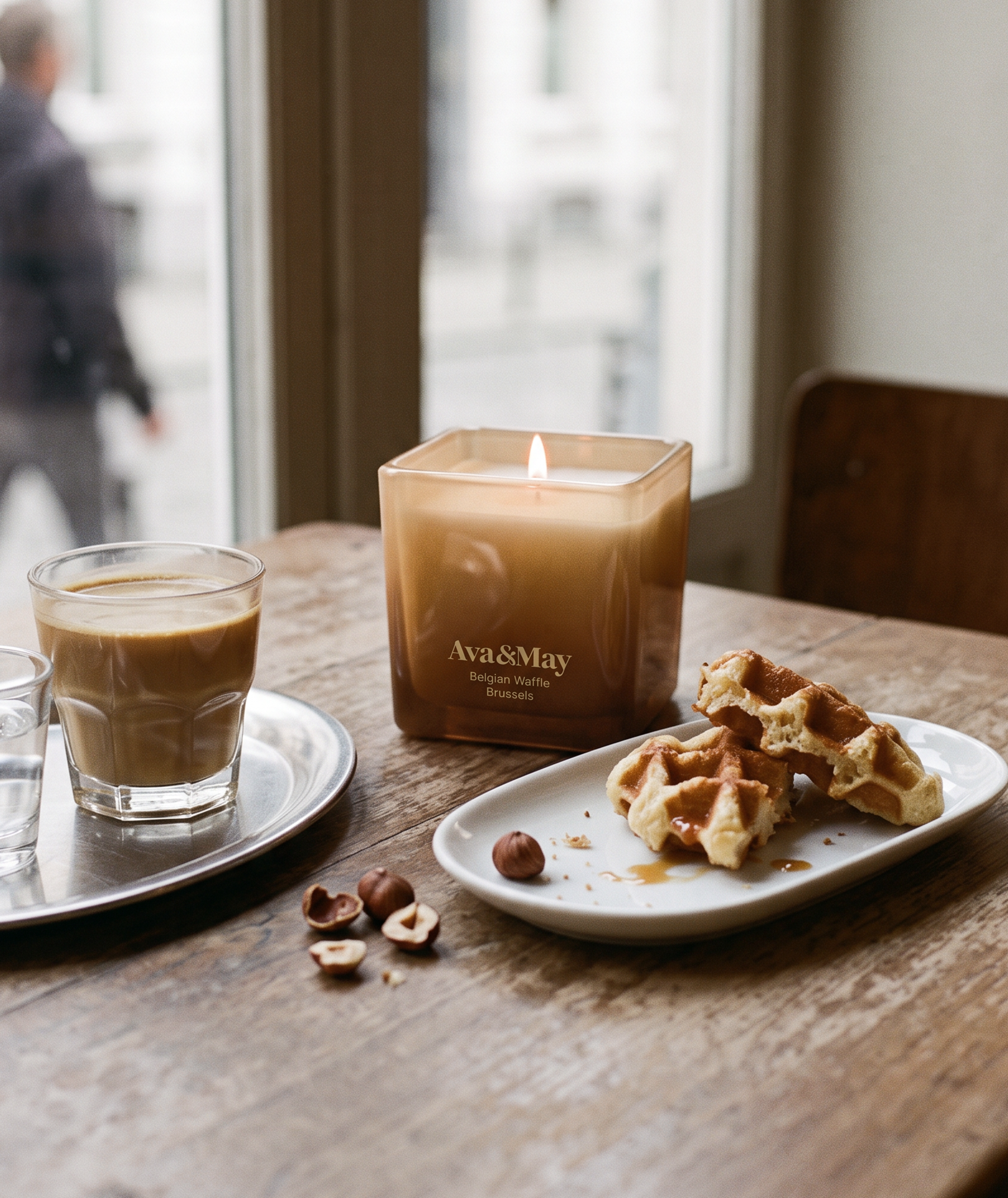 Bougie Ava&May Belgian Waffle de 220 grammes allumée dans un pot en verre carré, posée sur une table en bois à côté d'un verre de café, de noisettes et d'une gaufre belge sucrée