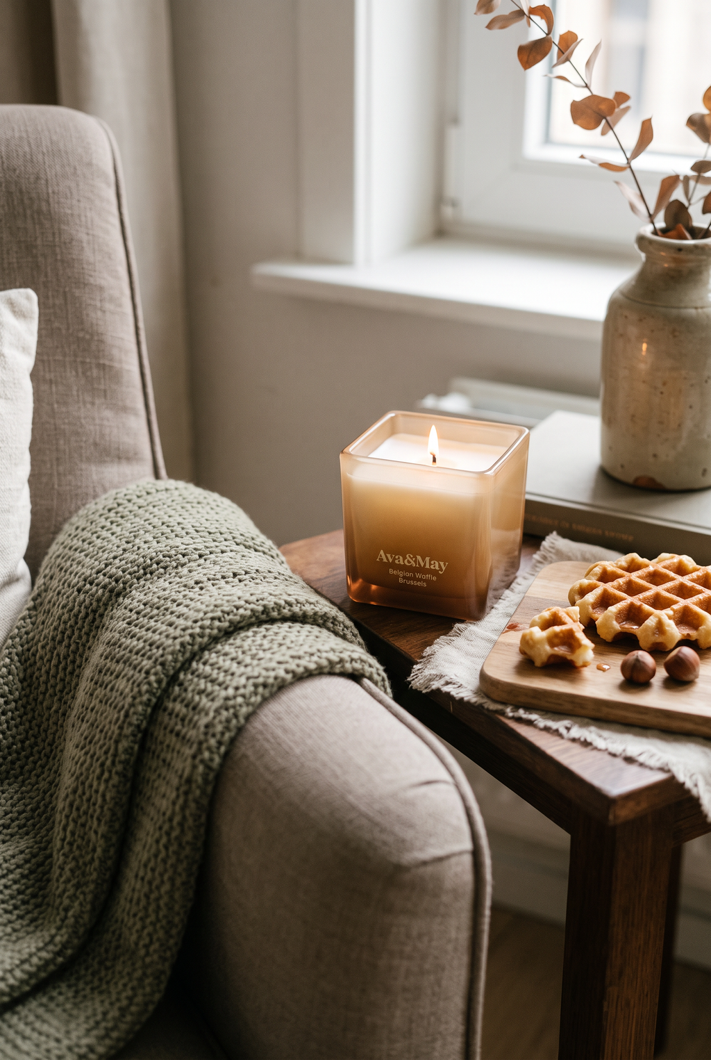La bougie Ava&May Belgian Waffle de 220 grammes, allumée, est posée sur une petite table en bois près d'un fauteuil beige recouvert d'une couverture en laine. À côté de la bougie se trouvent une gaufre belge et un vase de fleurs.