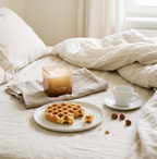 Bougie Ava&May Belgian Waffle de 220 grammes, allumée et posée sur un lit défait, avec une gaufre belge, une petite tasse de café et des noisettes