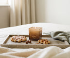 La bougie Ava&May Belgian Waffle de 220 grammes, allumée, est posée sur un plateau en bois, lui-même posé sur une nappe, entourée de petites gaufres et de noisettes.