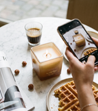 La bougie Ava&May Belgian Waffle de 220 grammes allumée sur une table en marbre, entourée d'un café expresso, d'une gaufre belge, d'un magazine de décoration d'intérieur et de noisettes, tandis qu'elle est photographiée par une personne tenant un téléphone à la main