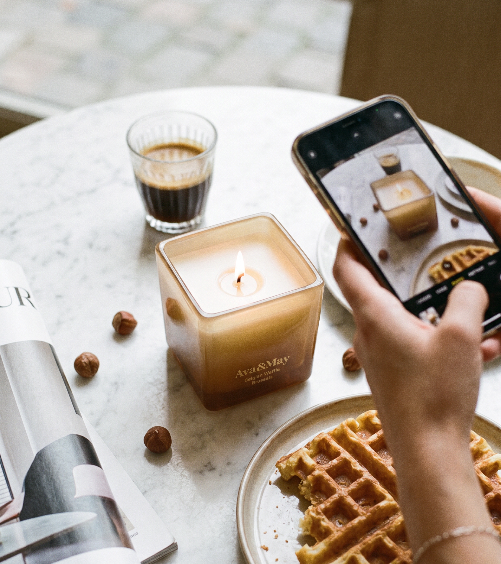 La bougie Ava&May Belgian Waffle de 220 grammes allumée sur une table en marbre, entourée d'un café expresso, d'une gaufre belge, d'un magazine de décoration d'intérieur et de noisettes, tandis qu'elle est photographiée par une personne tenant un téléphone à la main