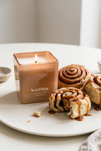 La bougie Cinnamon Roll de 220 grammes d'Ava&May, allumée sur une soucoupe en céramique, accompagnée de petits gâteaux à la cannelle épicés et nappés de crème au caramel