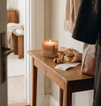 Une bougie Ava&May « Cinnamon Roll » allumée sur une petite table d'entrée en bois, accompagnée d'une pâtisserie à la cannelle