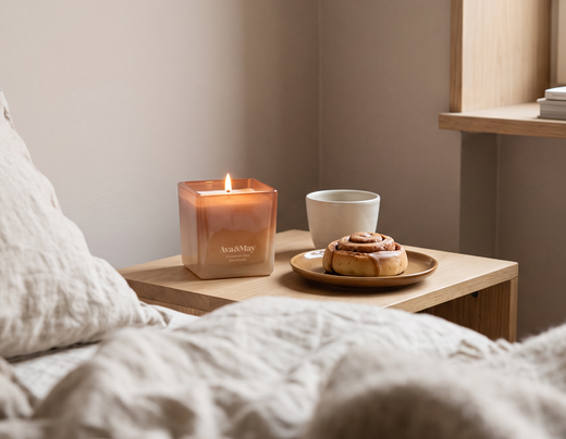 La bougie Cinnamon Roll de 220 grammes de la marque Ava&May, allumée sur une table de chevet en bois près d'un lit aux draps beiges. À côté se trouvent une petite pâtisserie à la cannelle et une tasse blanche.