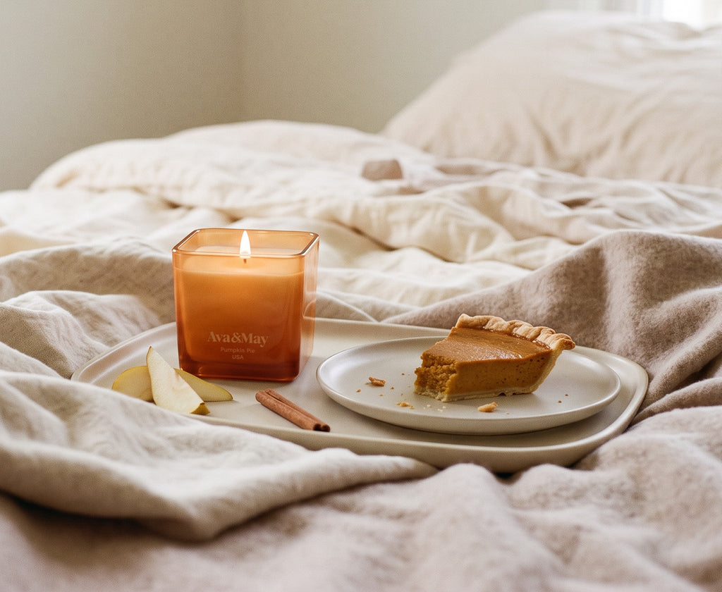 Une bougie Pumpkin Pie de 220 g de la marque Ava&May allumée sur un plateau posé sur un lit, accompagnée d'une part de tarte à la crème de potiron, aux poires et à la cannelle