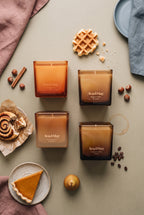 Coffret de bougies Sweet Sensations d'Ava&May : les bougies Belgian Waffle 220 grammes, Viennese Coffee 220 grammes, Pumpkin Pie 220 grammes et Cinnamon Roll 220 grammes sont posées sur une table verte, et à côté se trouvent un gâteau à la citrouille, un petit gâteau à la cannelle, de la cannelle, une gaufre belge, des noisettes et des grains de café