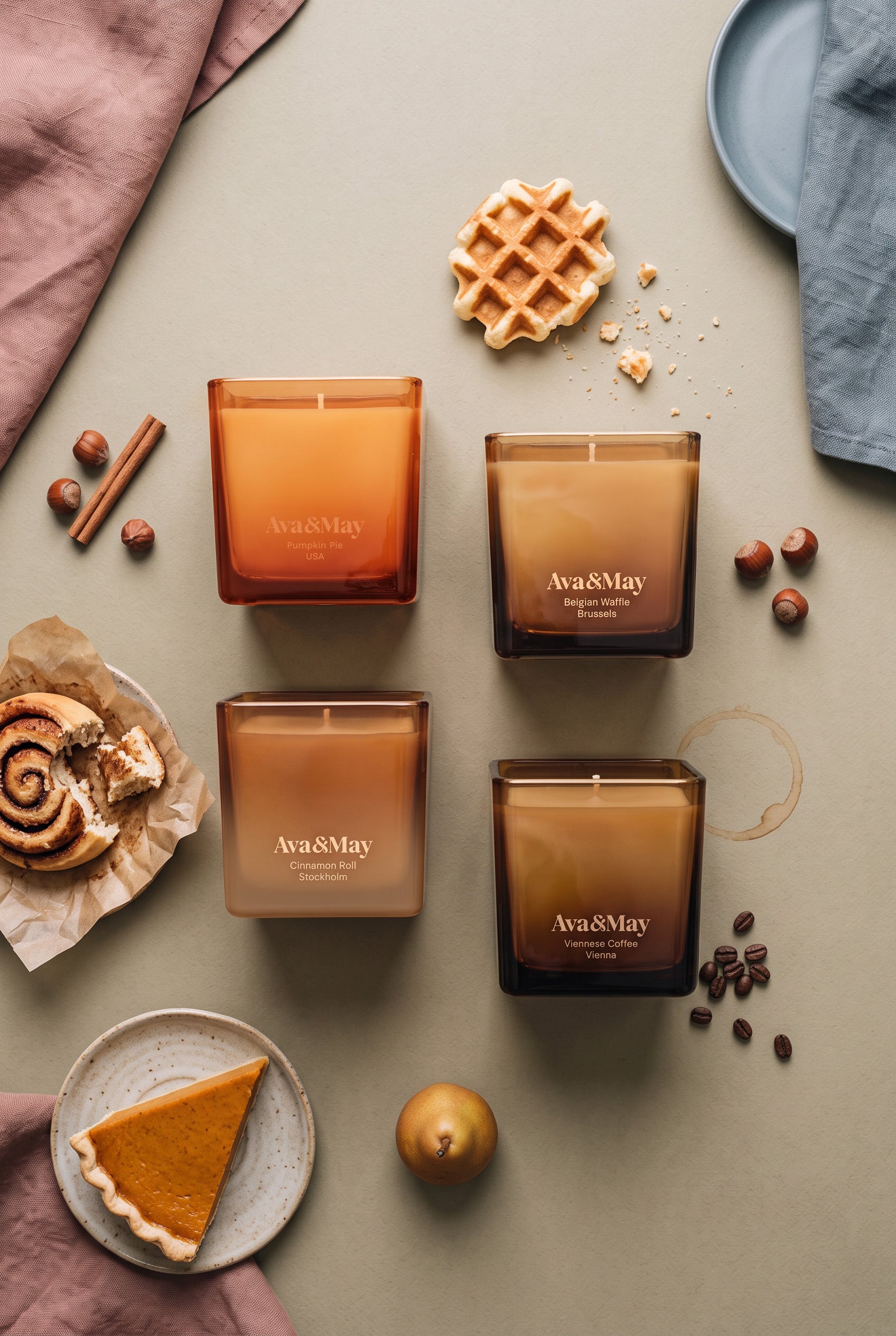 Coffret de bougies Sweet Sensations d'Ava&May : les bougies Belgian Waffle 220 grammes, Viennese Coffee 220 grammes, Pumpkin Pie 220 grammes et Cinnamon Roll 220 grammes sont posées sur une table verte, et à côté se trouvent un gâteau à la citrouille, un petit gâteau à la cannelle, de la cannelle, une gaufre belge, des noisettes et des grains de café