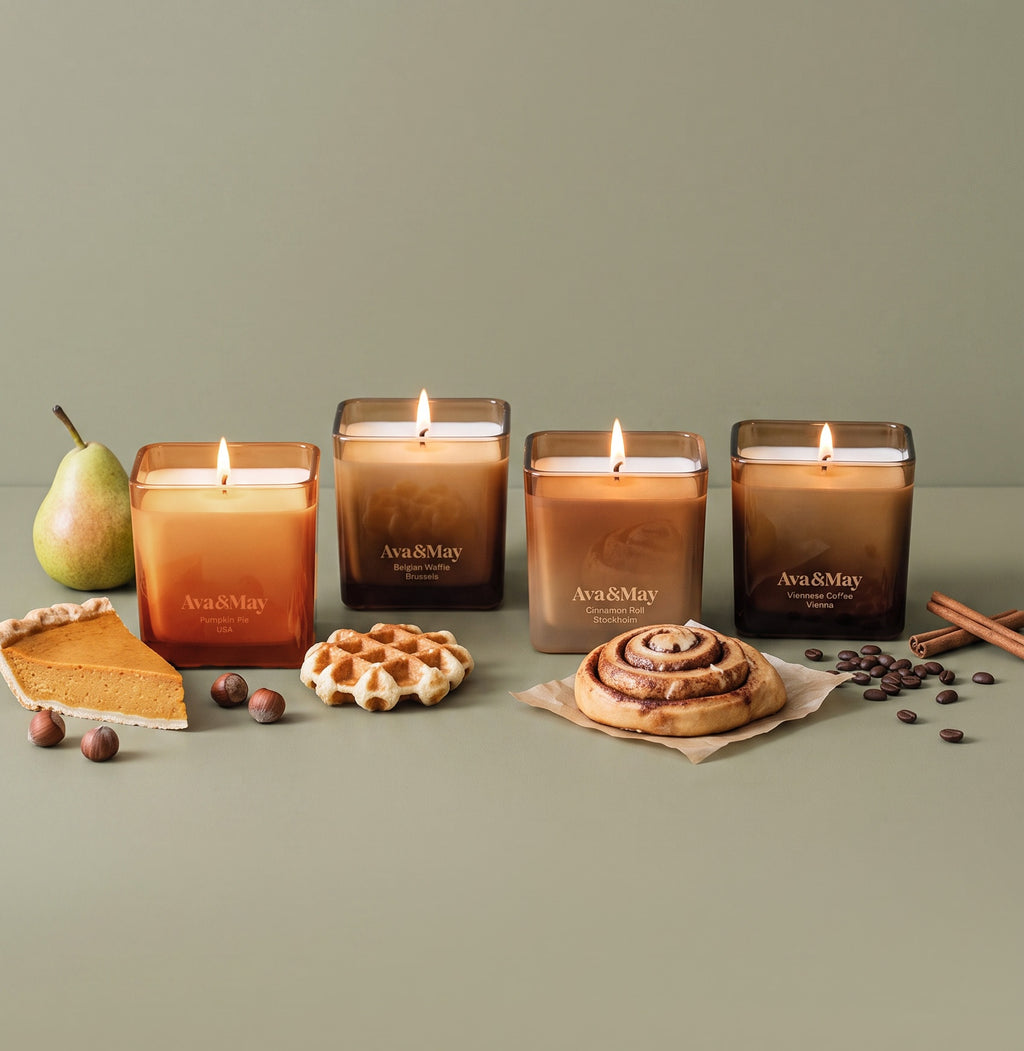 Coffret de bougies Sweet Sensations d'Ava&May : bougies Belgian Waffle 220 grammes, Viennese Coffee 220 grammes, Pumpkin Pie 220 grammes et Cinnamon Roll 220 grammes, allumées sur une table sont disposées en rangée et, à côté, se trouvent des petits gâteaux, une poire, des bâtons de cannelle, des grains de café et des noisettes
