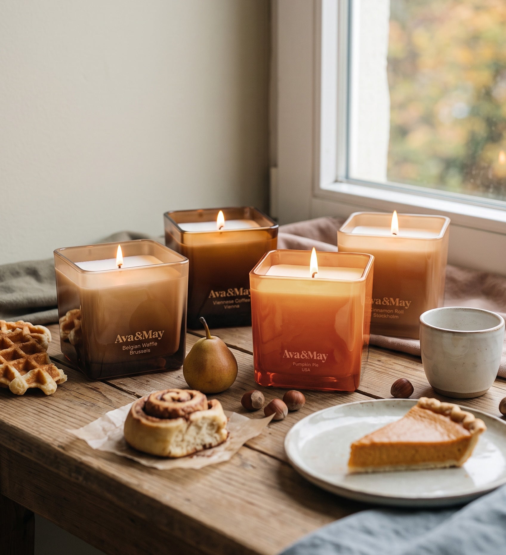 Coffret de bougies Sweet Sensations d'Ava&May : bougies Belgian Waffle 220 grammes, Viennese Coffee 220 grammes, Pumpkin Pie 220 grammes et Cinnamon Roll 220 grammes, allumées sur une table en bois où sont disposées des gaufres, une tarte à la citrouille et des brioches à la cannelle